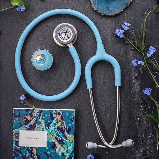 Littmann Classic III Monitoring Stethoscope: Satin Marine Blue Tube 5912C, notebook, candle, and small blue flowers on a dark textured background.