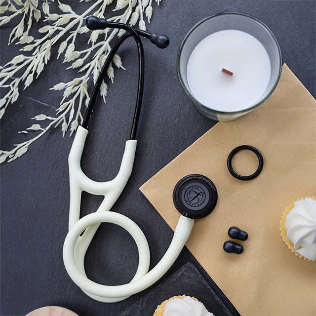 A 3M™ Littmann® Cardiology IV Diagnostic Stethoscope: Satin Alabaster Tube 6186C, a candle, and black pills arranged around white cupcakes on a dark textured surface.