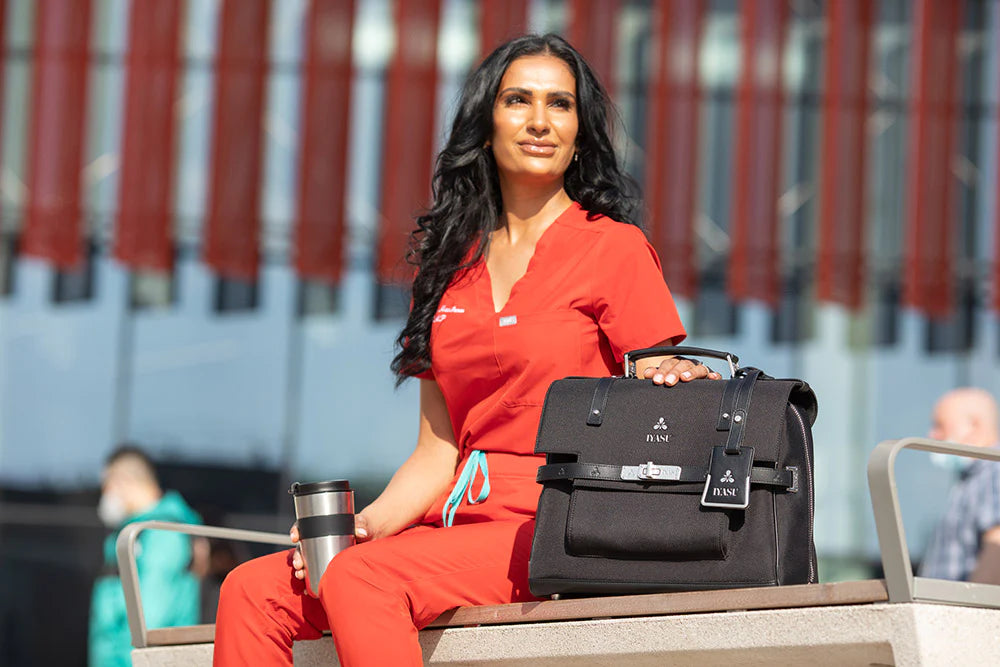 A woman in red scrubs sitting on a bench holding an IYASU Virginia Medical Bag In Black.