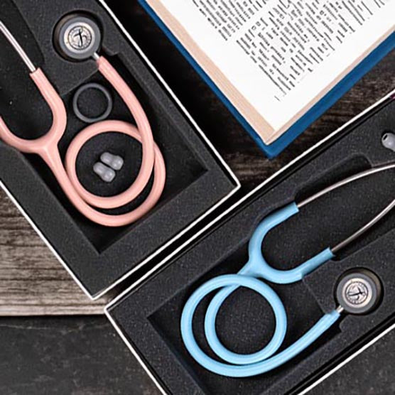 Two open boxes displaying a pink and a blue 3M™ Littmann® Classic III Monitoring Stethoscope: Satin Champagne Rose Tube 5910C, featuring dual tunable diaphragms, next to an open book on a wooden surface.